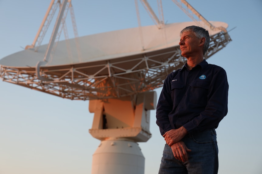 A man stands and looks into the distance. Behind him is a large white telescope.