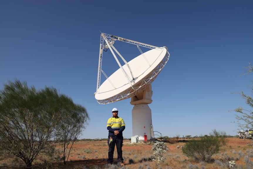 A man is standing in the desert with a large telescope behind him.