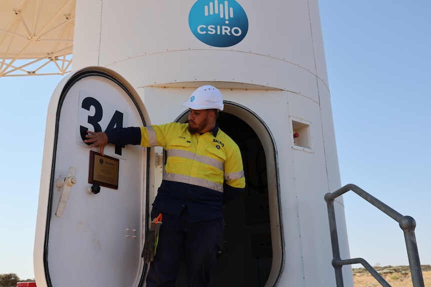 A man standing by a telescope looking at a plaque with his hand on it.